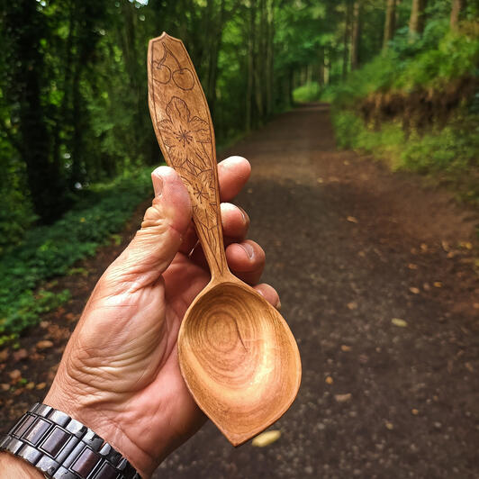 kolrosed cherry spoon A cherry spoon rests in the hand with a pathed woodland in the background. The spoon has a detailed cherry blossom design kolrosed onto the handle to further embellish the story of the spoons origin