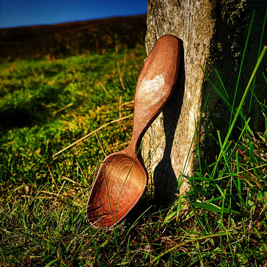 baked alder eater A baked alder symmetrical eating spoon rests in the golden sunlight against the base of a stone wall with a beautiful summer meadow as a backdrop