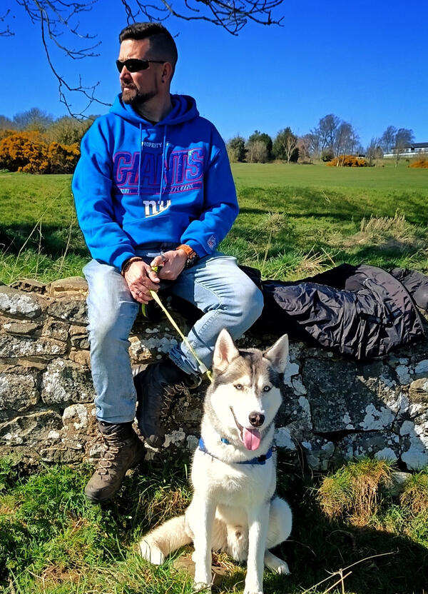 Nick Rowe profile pic Nick sitting on a wooden bench by a stone wall looking into the distance with content