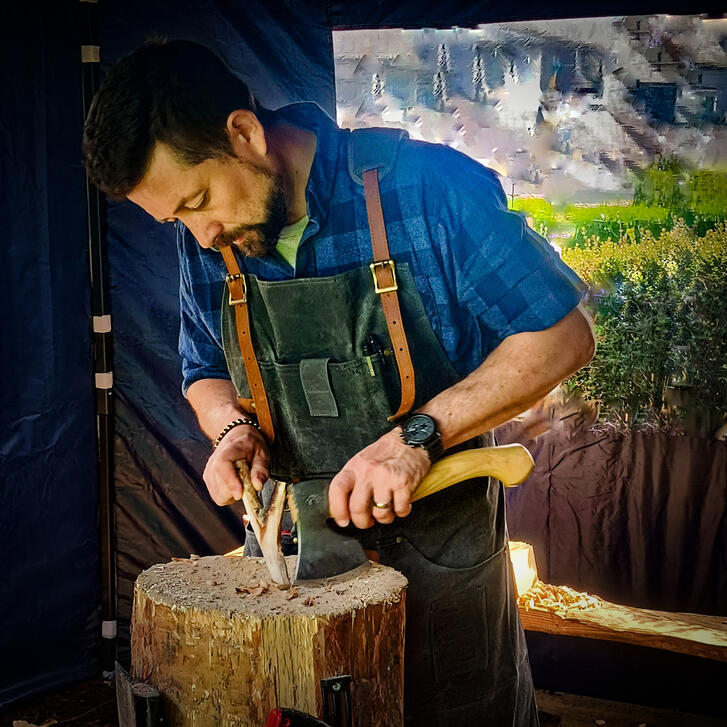 hard at it The maker Nick working on piece in his workspace, standing at the chopping block with axe, green canvas apron and blue checked shirt